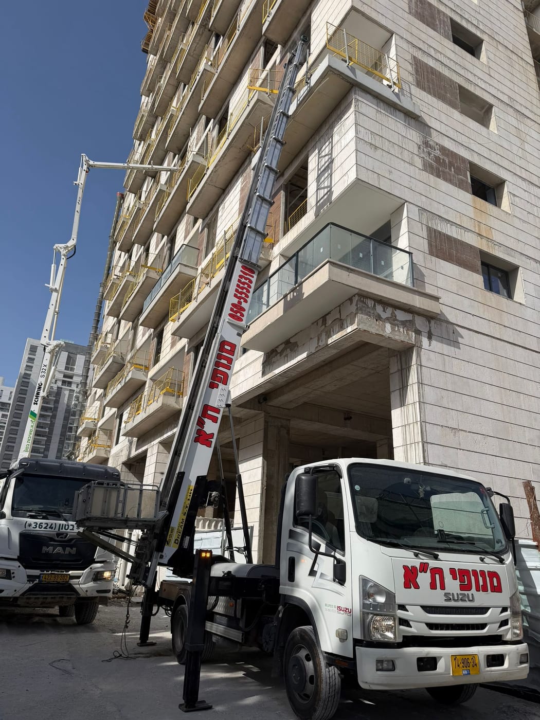 מנוף סולם פרוס לאורך חזית בניין בשלבי בנייה עם מעקות בטיחות צהובים, לצד זרוע משאבת בטון באתר