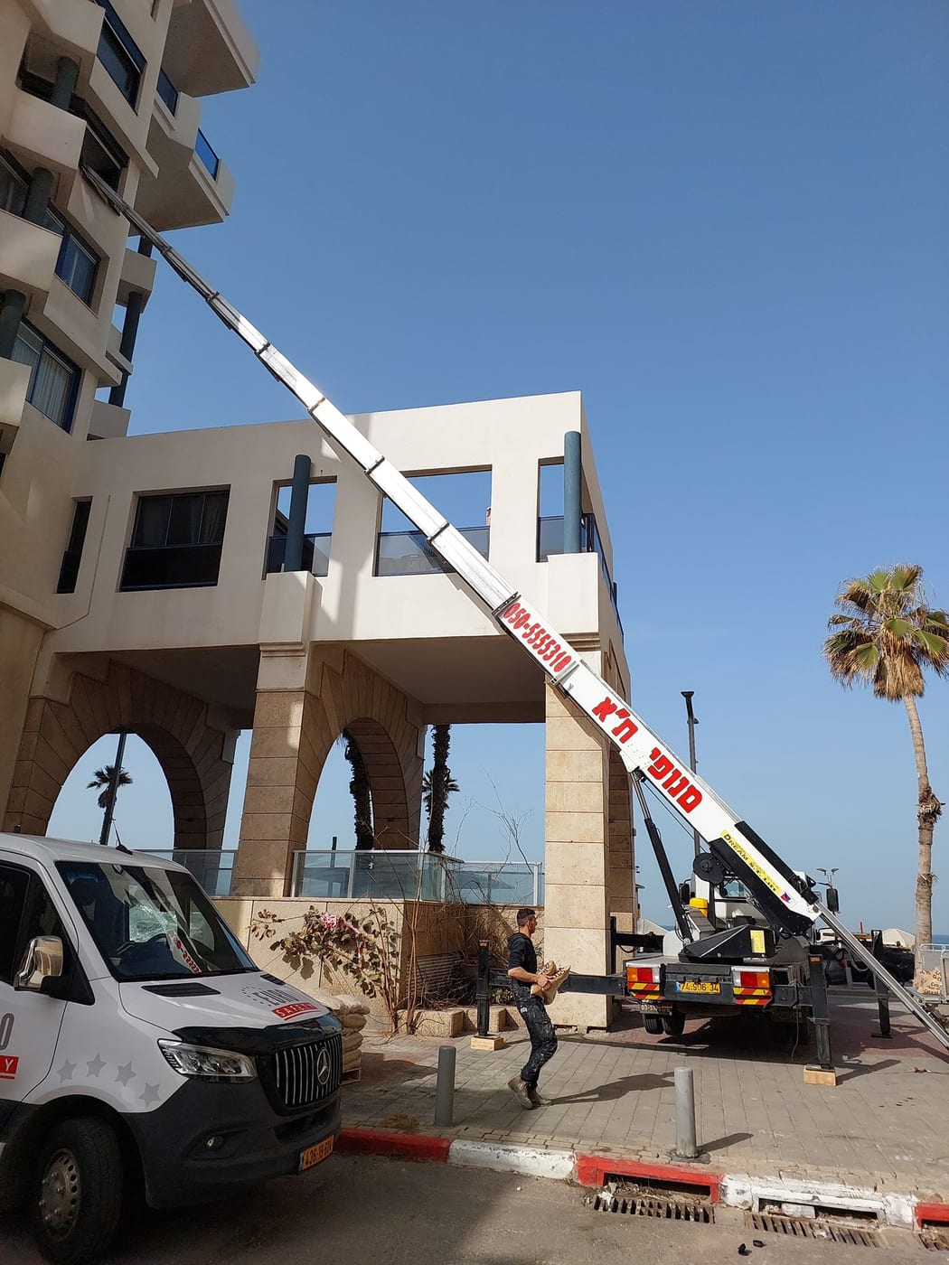מנוף סולם ממותג מנופי תל אביב פרוס אל קומה גבוהה בבניין בקו הראשון לים, עם קולונדה מקושתת ועצי דקל ברקע
