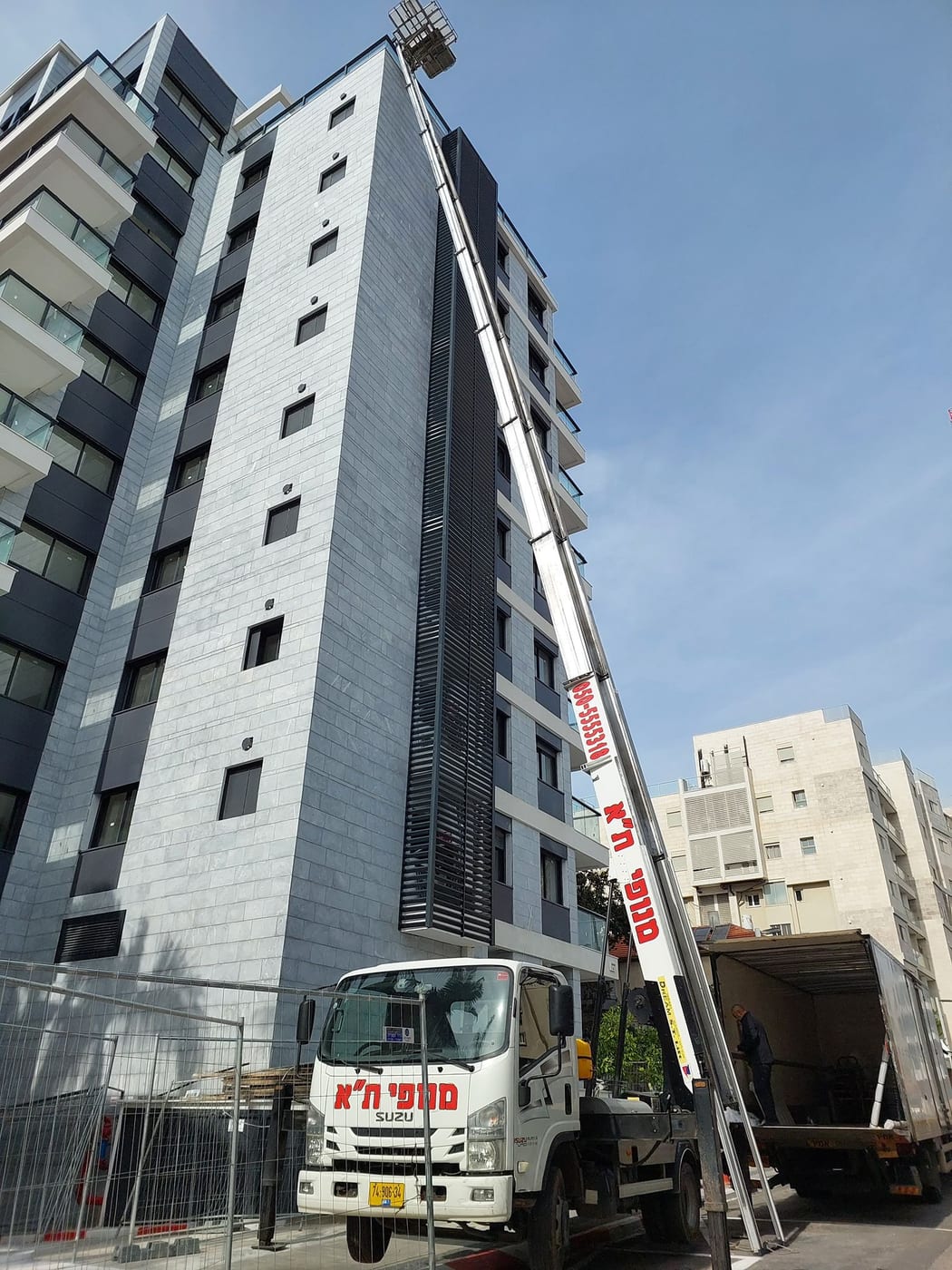 מנוף סולם של מנופי תל אביב פרוס במלוא אורכו עד גג מגדל מגורים מחופה אבן אפורה, כשמשאית ההובלה עומדת לידו