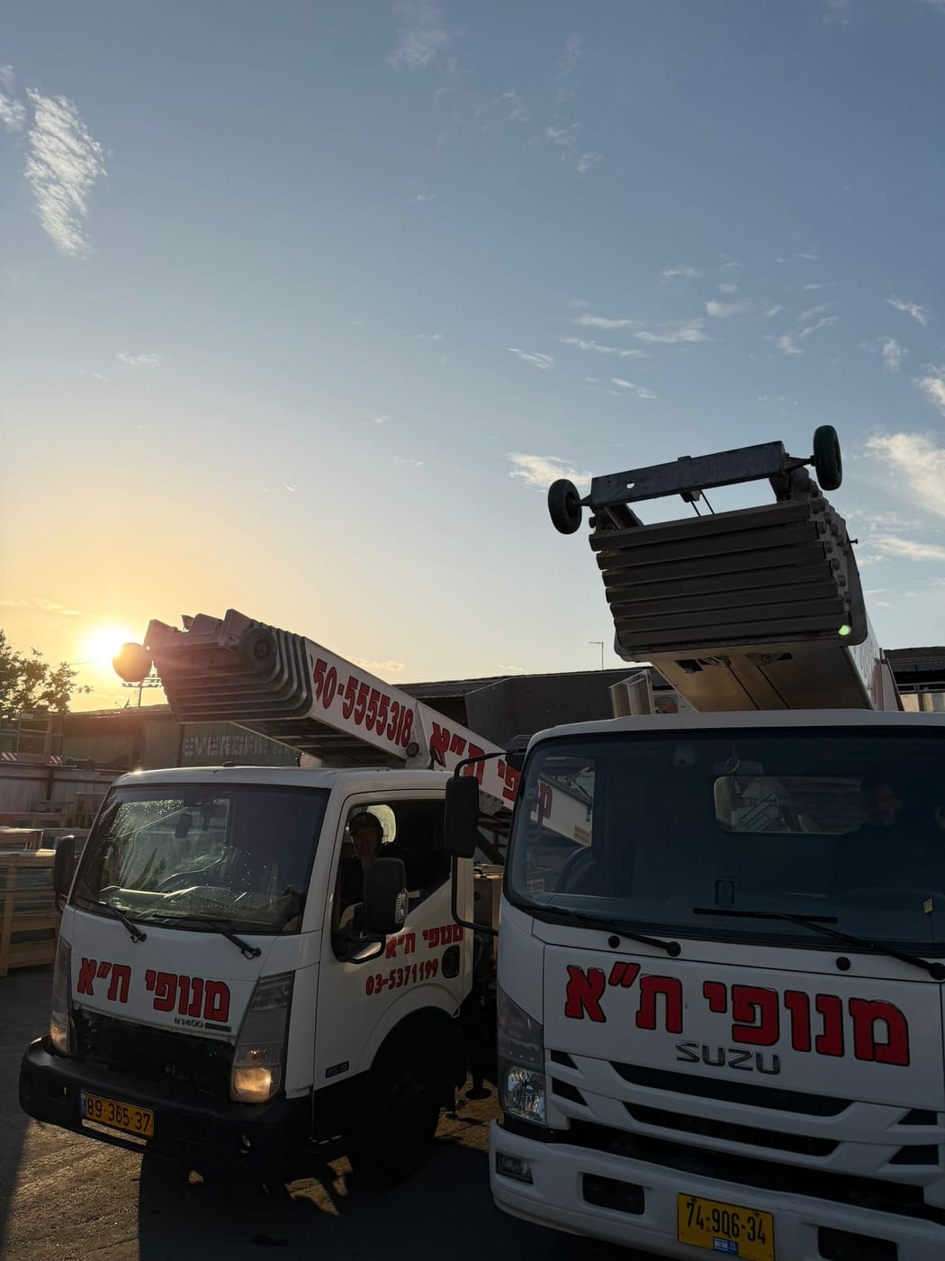 שתי משאיות מנוף סולם ממותגות מנופי ת"א בחצר החברה בשעת שקיעה, הזרועות מקופלות