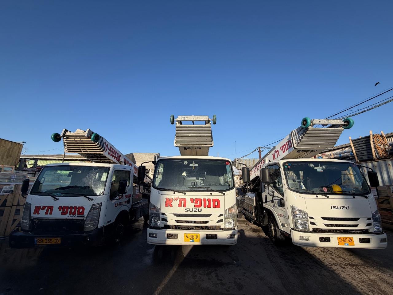 שלוש משאיות מנוף סולם ממותגות מנופי ת"א חונות בחצר החברה, הזרועות מקופלות, שמיים כחולים